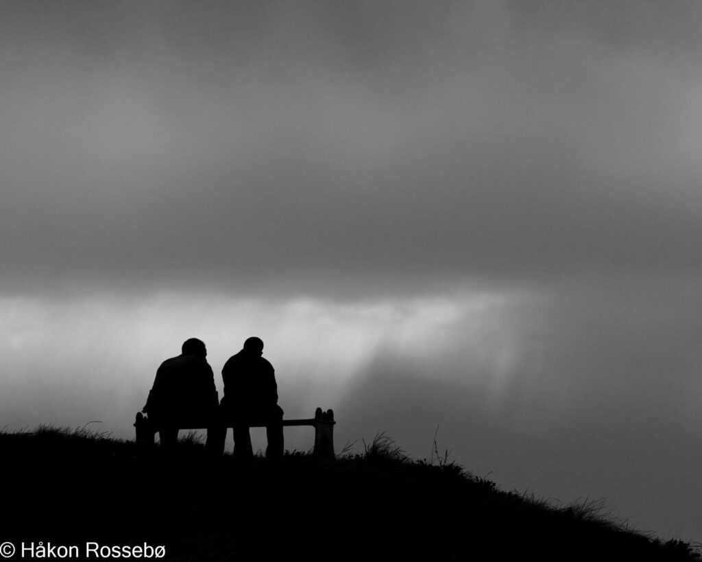 Silhuett av to menn i samtale på benk, mørke skyer